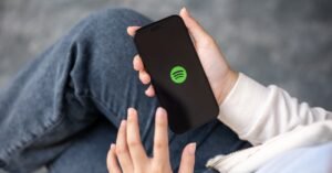 Person in white hoodie and blue jeans holding a smartphone with Spotify app icon on screen, against a blurred gray background.