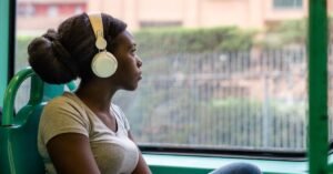 Person with hair in a bun wearing white headphones and a gray shirt, sitting on a bus and looking out the window with a green frame and urban background.