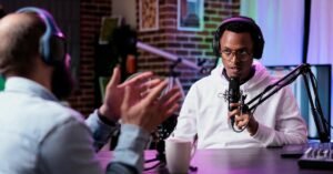 Person clapping during a podcast interview, other speaking into a microphone in a studio with brick wall.