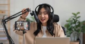 Podcaster in a home studio recording with a microphone, headphones, and smartphone camera, surrounded by plants and shelves.
