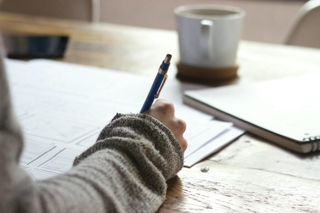 How to Write a Podcast Script? Person writing with a blue pen on paper at a wooden desk.